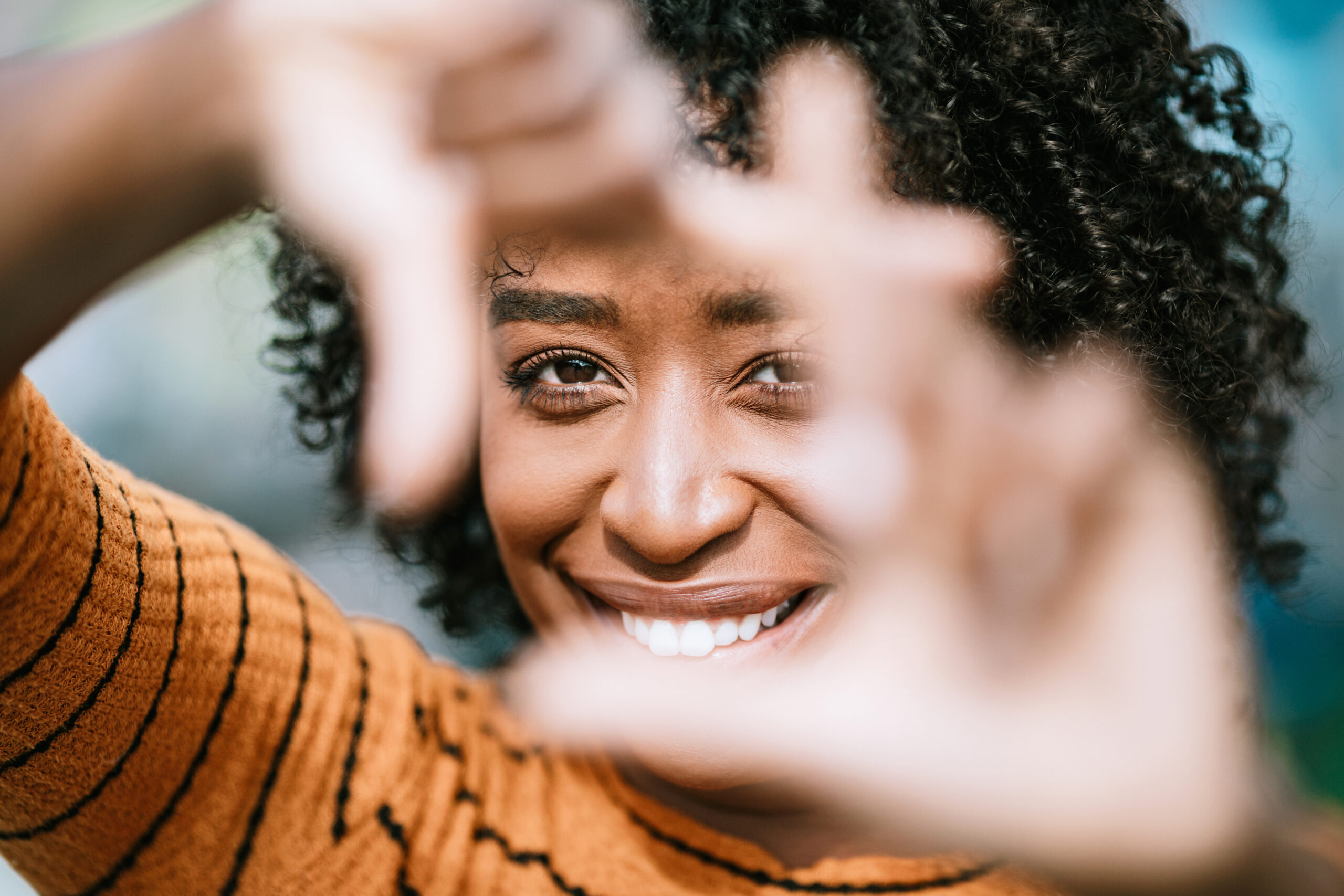 Natural Beauty Portrait With Womans Finger Frame Eine lächelnde Frau mit dunkler, lockiger Frisur trägt einen orangefarbenen Pullover mit schwarzen Streifen. Die Person hält beide Hände vor das Gesicht und formt mit den Fingern einen rechteckigen Rahmen. Der Hintergrund ist unscharf und zeigt angedeutete Farben. Das Bild stellt symbolisch den Mensch im Mittelpunkt dar.