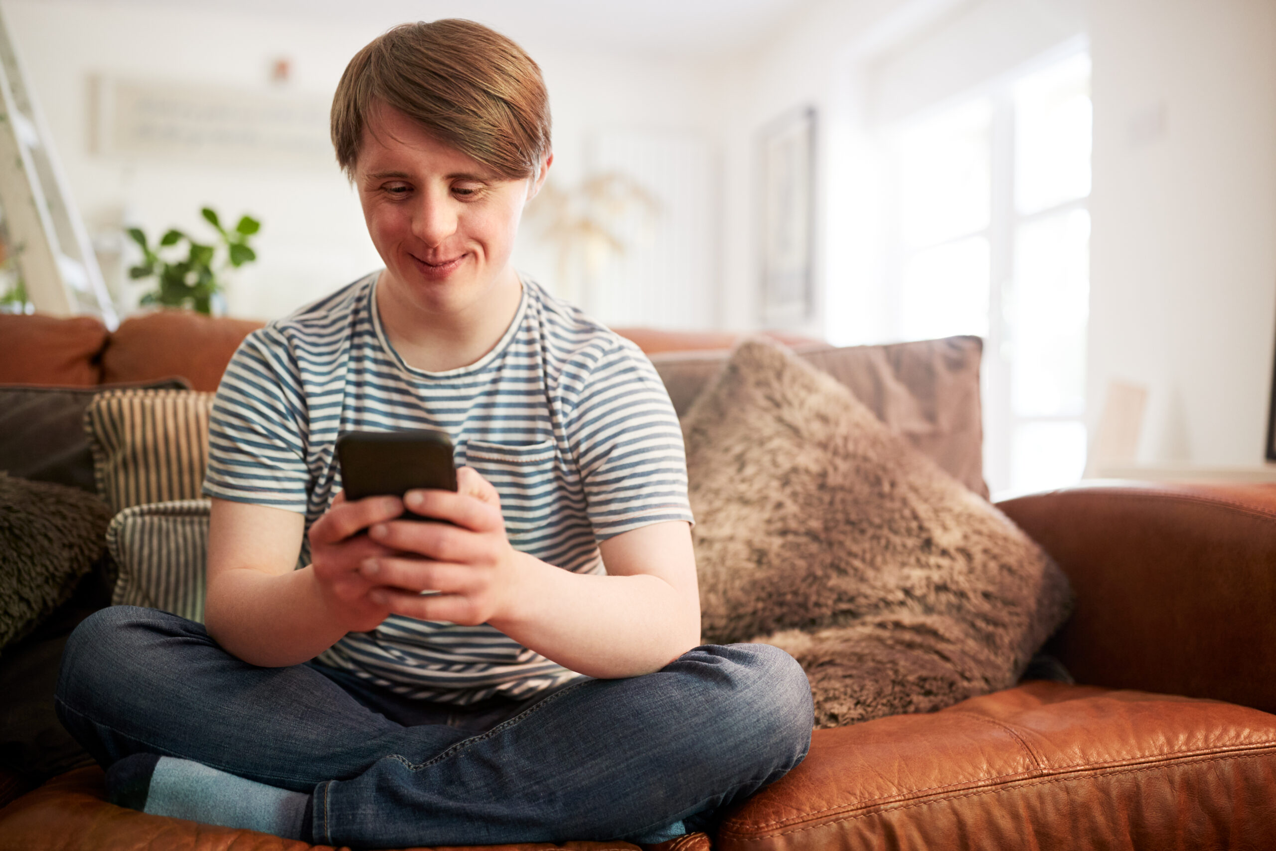 Young Downs Syndrome Man Sitting On Sofa Using Mobile Phone At Home Ein junger Mann sitzt auf einem braunen Ledersofa. Er hat die Beine übereinander gekreuzt. Er trägt ein kurzärmeliges, gestreiftes T-Shirt und eine blaue Jeans. Er hält ein Smartphone mit beiden Händen und schaut darauf. Im Hintergrund sind große Kissen, Pflanzen und ein helles Fenster zu sehen. Die Szene wirkt freundlich. Das Bild symbolisiert, dass der junge Mann selbst die Entscheidung trifft, wie er Unterstützung und Hilfen bekommt, und damit die Kontrolle über sein Leben hat.
