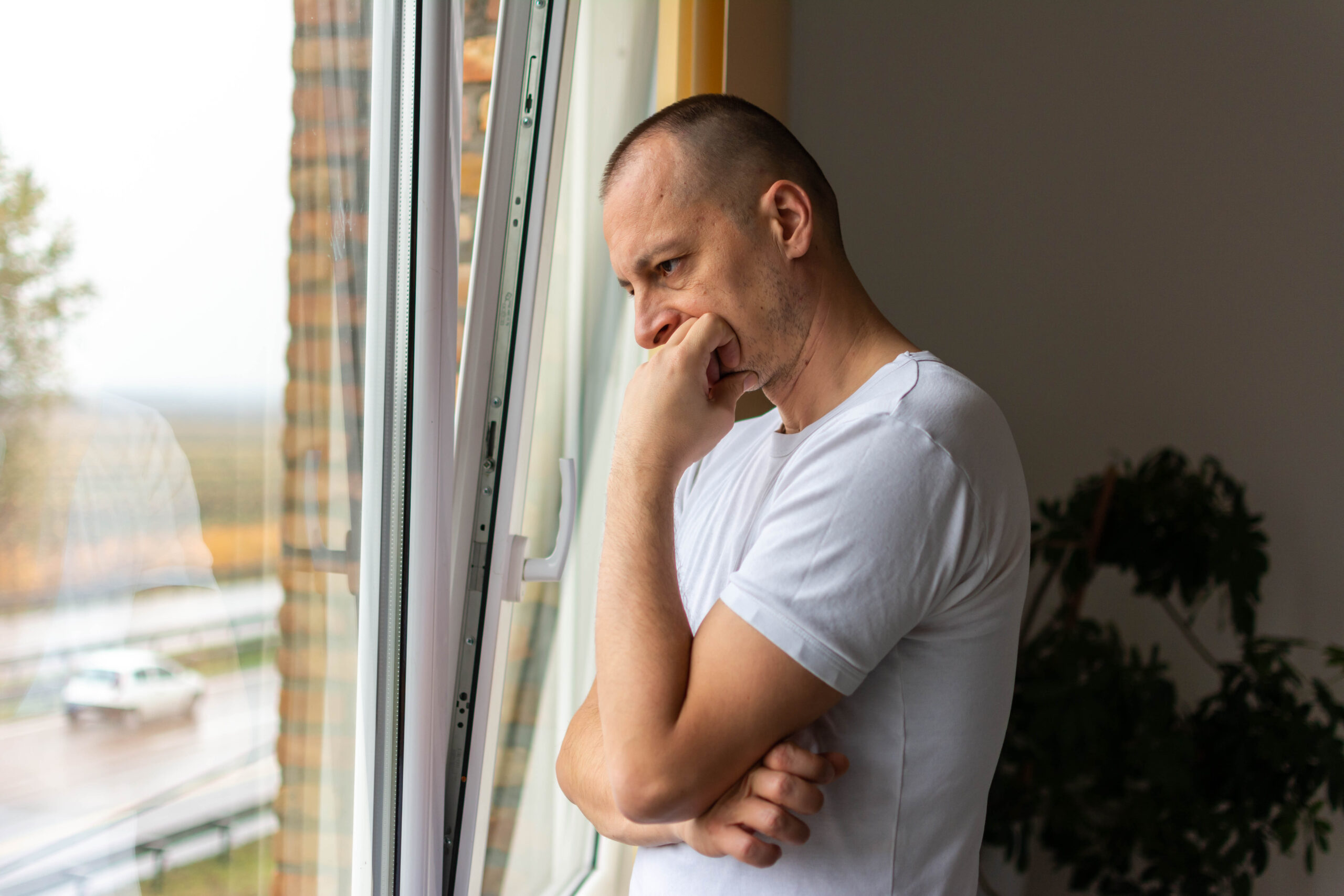 thoughtful entrepreneur leaning against a glass window in his office Ein Mann steht in einem hellen Raum an einem geöffneten Fenster und blickt nach draußen. Er trägt ein weißes T-Shirt und stützt den Kopf nachdenklich auf eine Hand. Im Hintergrund ist eine grüne Zimmerpflanze zu sehen, draußen erkennt man eine Straße mit vorbeifahrendem Auto und unscharfer Landschaft. Die Szene beschreibt einen einsamen depressiven Mann am Fenster, der sich hoffnungslos fühlt. Das Bild steht symbolisch für Menschen mit psychischer Erkrankung.