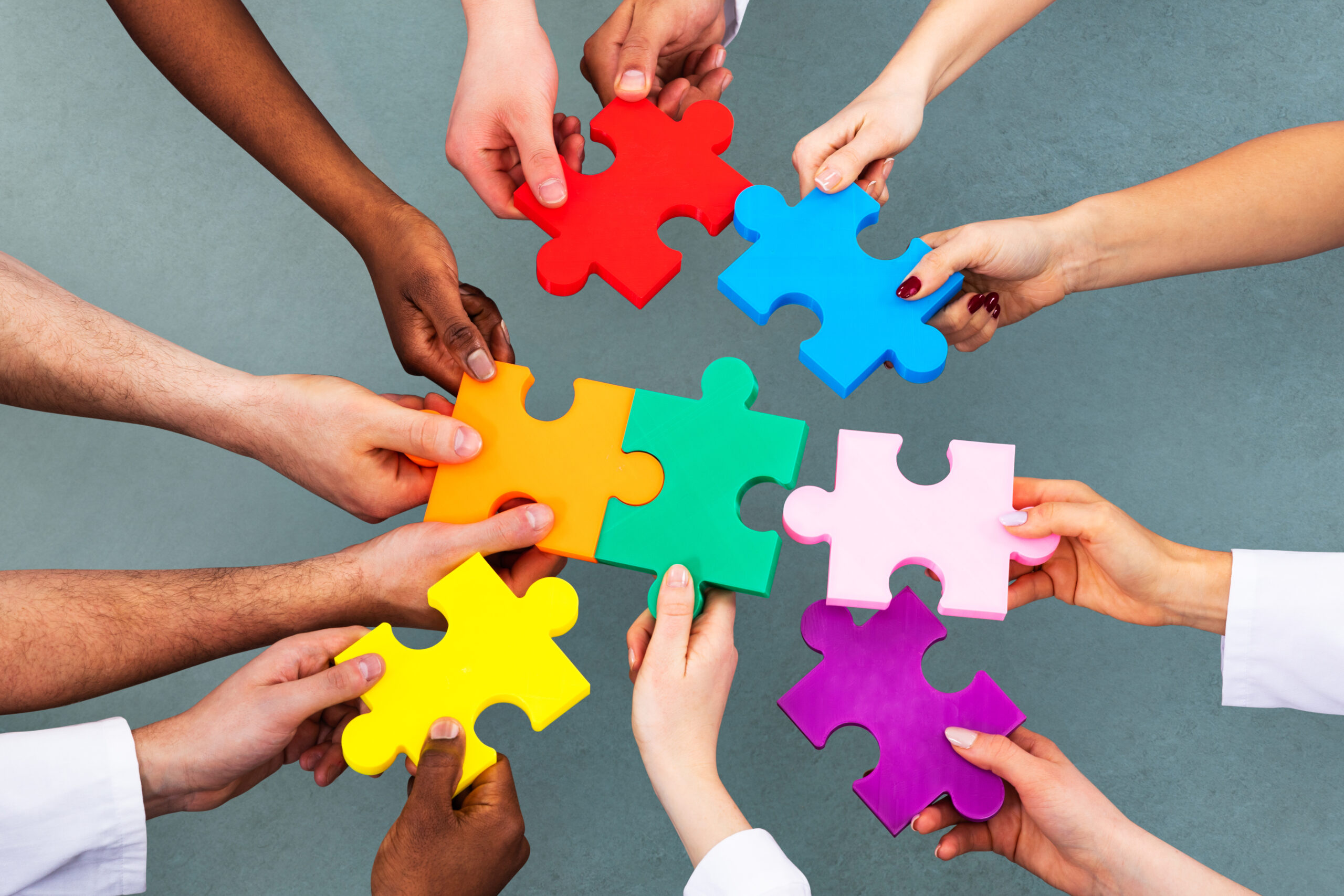 Zusammenarbeit und Vielfalt Das Bild zeigt mehrere Hände von Menschen mit unterschiedlicher Hautfarbe. Sie halten bunte Puzzleteile und fügen diese in der Mitte zusammen. Die Puzzleteile sind in verschiedenen Farben wie Rot, Blau, Grün, Gelb, Orange, Rosa und Lila. Der Hintergrund ist einfarbig und grau. Das Bild symbolisiert Zusammenarbeit und Vielfalt.
