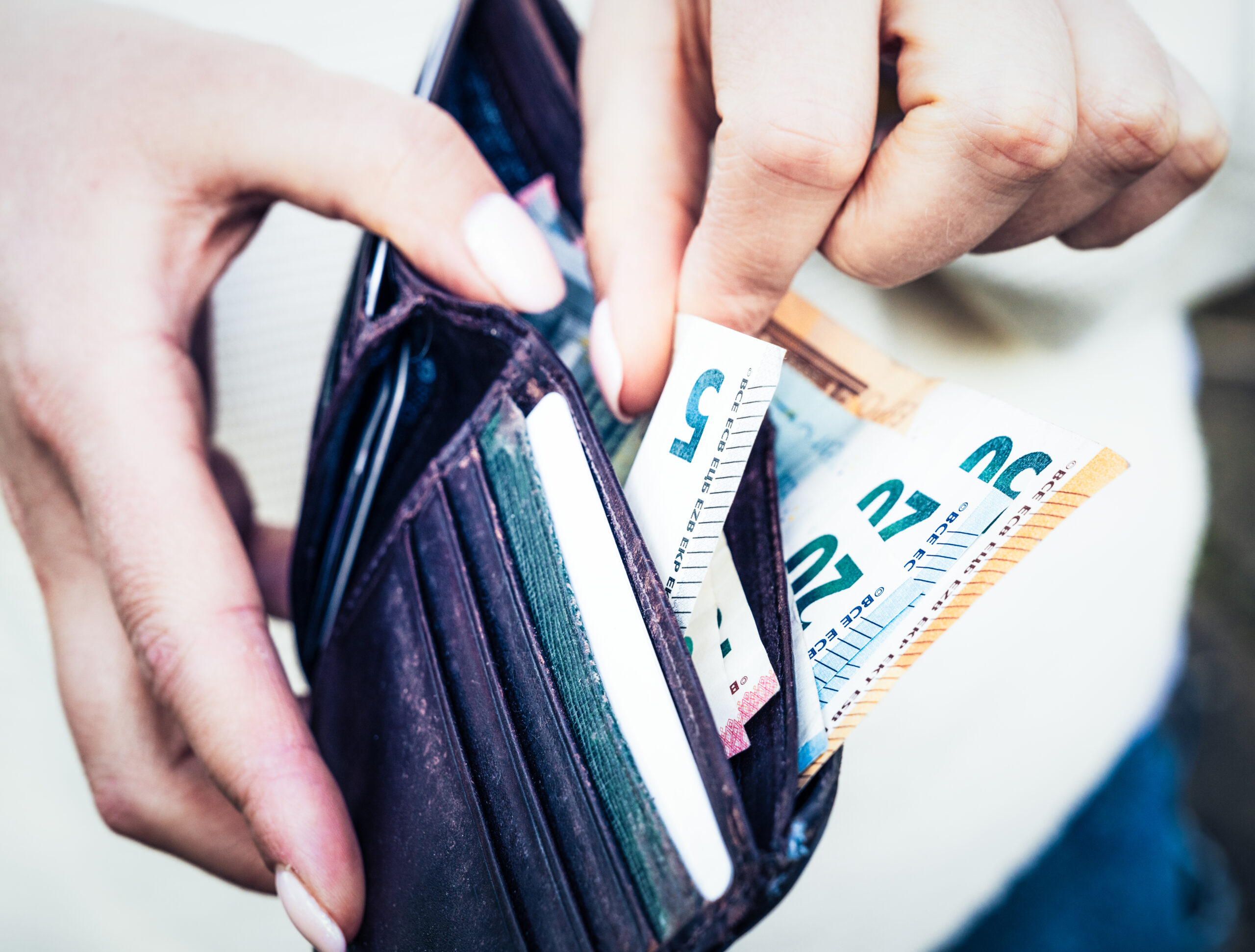 Taking cash from a wallet Eine Person hält eine geöffnete, dunkelbraune Geldbörse in den Händen. In der Geldbörse befinden sich mehrere Euro-Geldscheine, darunter ein Fünf-, ein Zwanzig- und ein Fünfzig-Euro-Schein, die teilweise herausragen. Außerdem sind Fächer mit Karten sichtbar. Die Szene ist nah aufgenommen und zeigt nur die Hände und die Geldbörse. Das Bild symbolisiert das selbstständige Bezahlen.