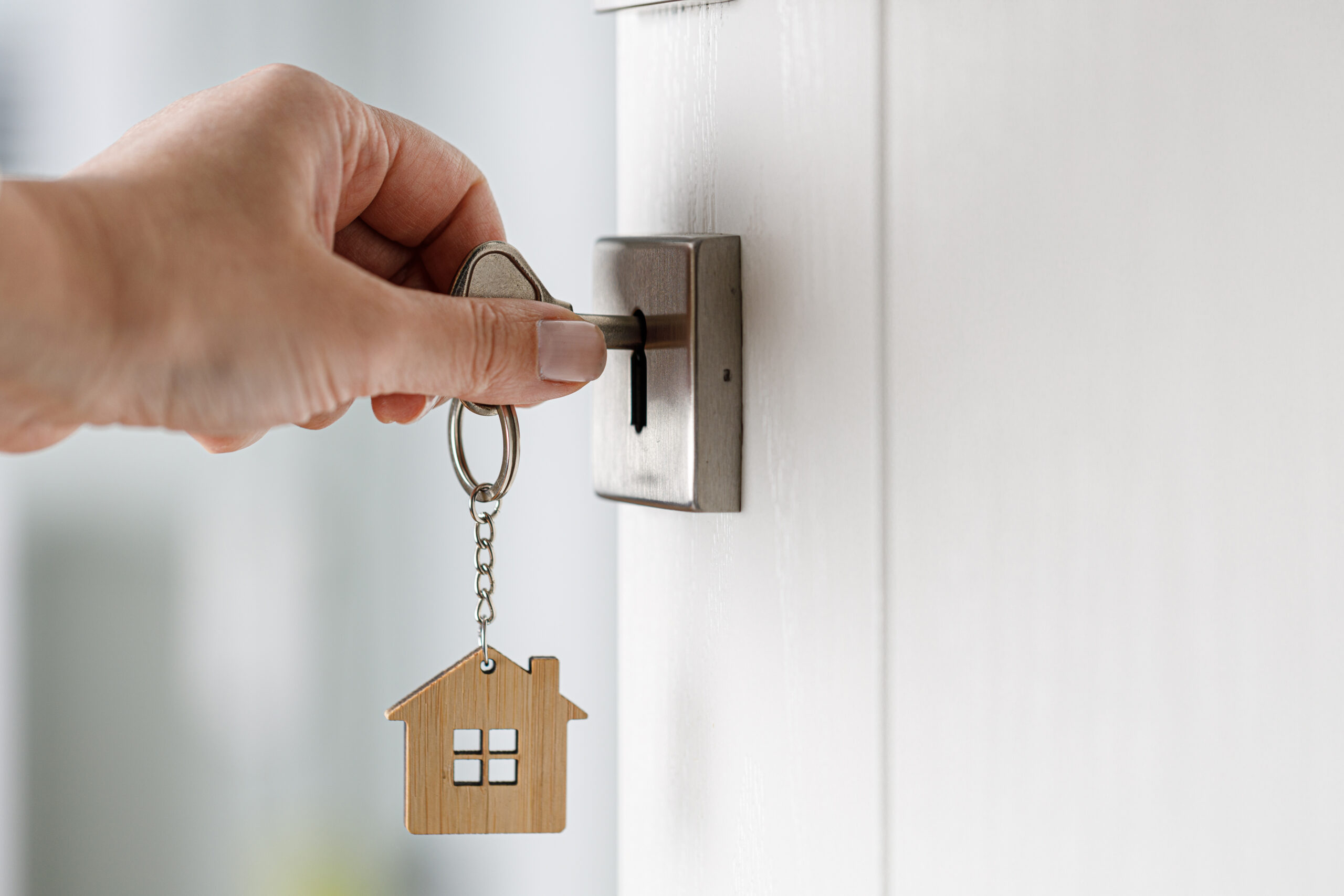 Die eigene Wohnung Eine Hand steckt einen Schlüssel in ein Türschloss. Am Schlüsselbund hängt ein Schlüsselanhänger in Form eines kleinen Hauses mit Fenstern. Die Tür ist weiß und das Schloss aus Metall. Der Hintergrund ist unscharf und hell. Dieses Bild soll die eigene Wohnung symbolisch darstellen.