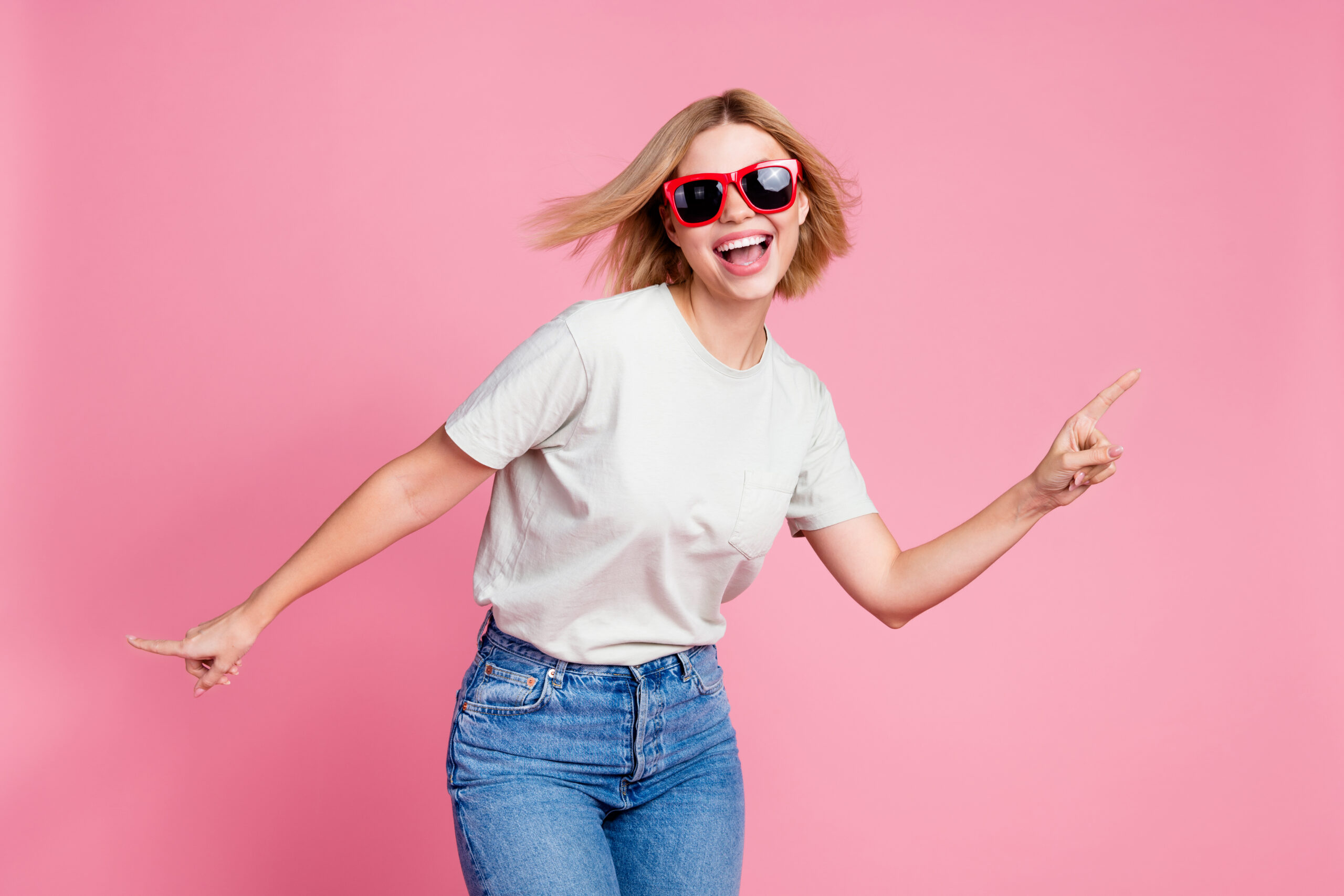 Photo of cheerful lovely girl wear stylish grey clothes dance have fun isolated on pink color background Das Bild zeigt eine junge Frau mit schulterlangem, blondem Haar. Sie trägt ein weißes T-Shirt und eine blaue Jeans. Im Hintergrund ist eine einfarbige, rosa Wand. Die Frau steht in einer lebendigen, tanzähnlichen Haltung. Sie hat die Arme ausgestreckt und zeigt mit den Zeigefingern in verschiedene Richtungen. Die Szene soll zeigen, dass die Frau selbst entscheiden kann und ihre eigene Stärke hat.