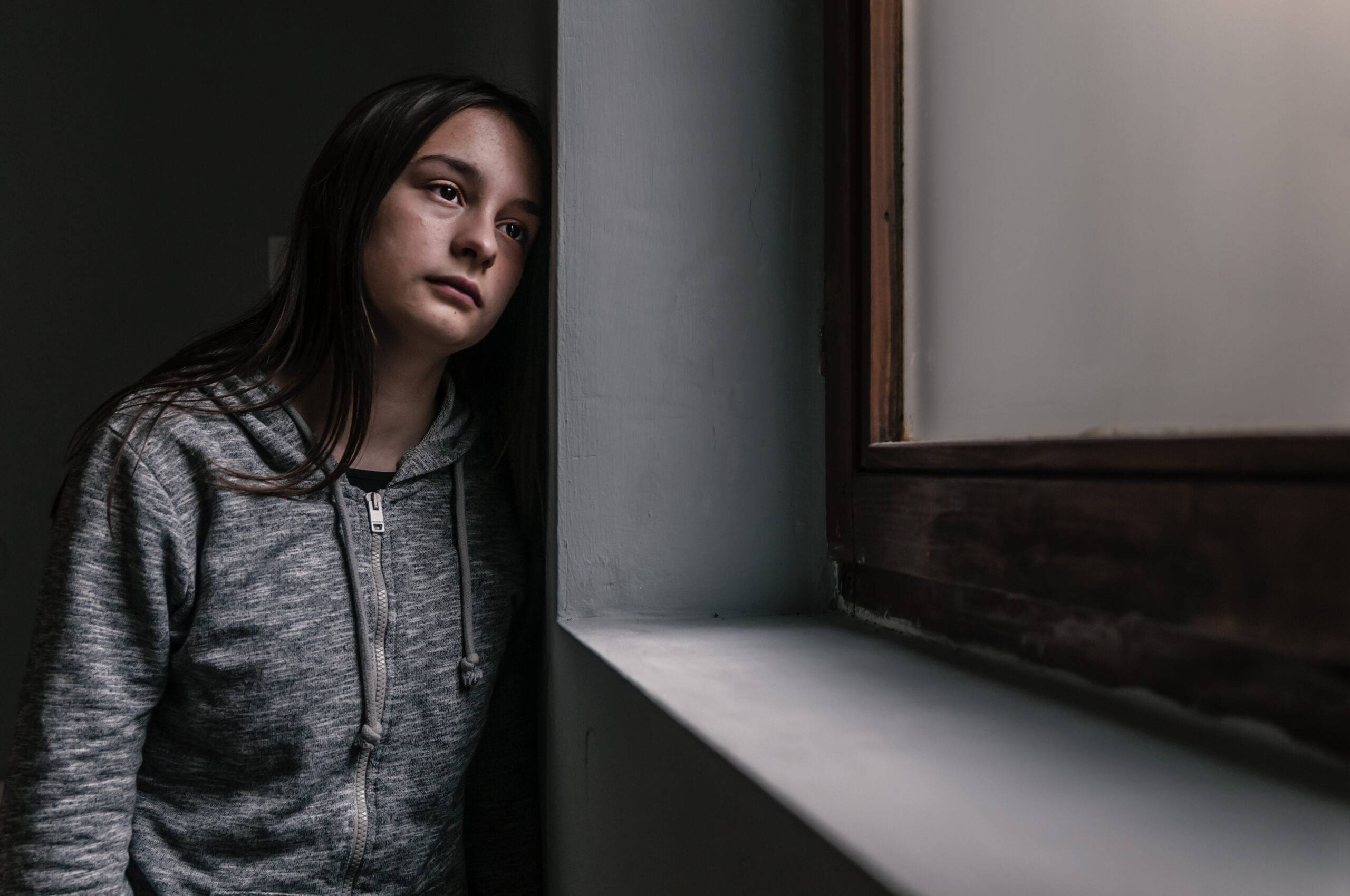 Sad Young Girl Eine junge Frau mit langen braunen Haaren steht in einem dunklen Raum vor einem Fenster. Sie trägt eine graue Kapuzenjacke mit Reißverschluss. Das Fenster befindet sich rechts im Bild und hat einen Rahmen aus Holz. Die Frau lehnt sich leicht nach vorne und schaut traurig nach draußen. Die Szene wirkt dunkel und nachdenklich. Das Bild soll symbolisieren, wie sich Menschen mit psychischen Beeinträchtigungen fühlen können.