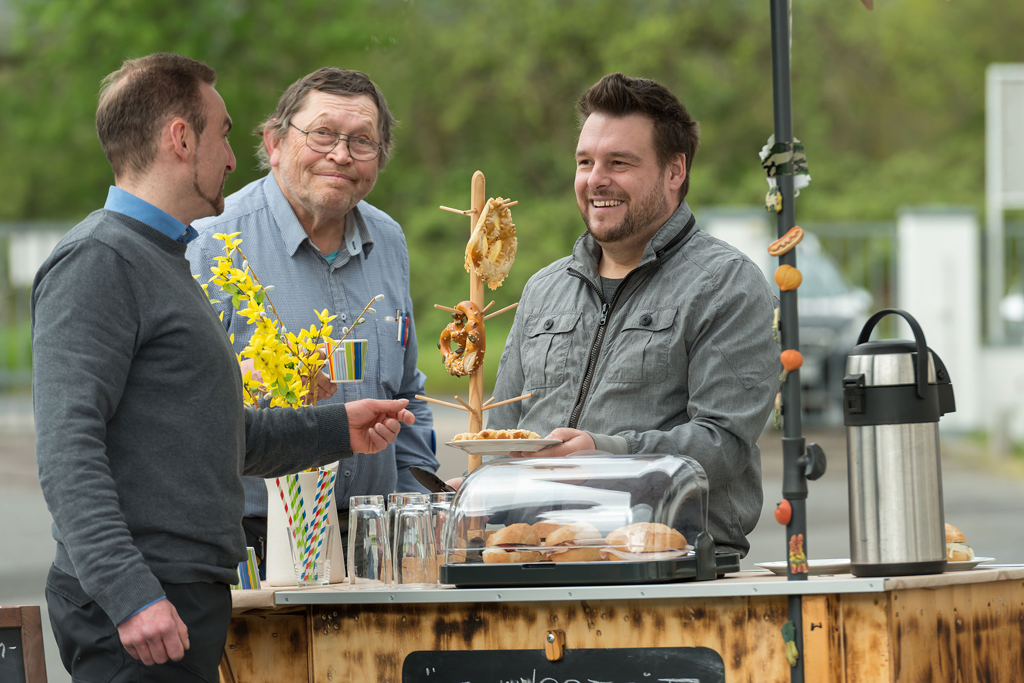 An einem mobilen Kiosk treffen sich drei Männer zu einer freundlichen Begegnung. Der jüngere Mann in grauer Jacke übergibt eine Käsebrezel. Der Stand ist mit einer Buffetvitrine, Getränken, einer Auswahl an belegten Brötchen sowie Brezeln ausgestattet. Die Umgebung deutet auf eine entspannte Pausenatmosphäre hin. An einem mobilen Kiosk treffen sich drei Männer zu einer freundlichen Begegnung. Der jüngere Mann in grauer Jacke übergibt eine Käsebrezel. Der Stand ist mit einer Buffetvitrine, Getränken, einer Auswahl an belegten Brötchen sowie Brezeln ausgestattet. Die Umgebung deutet auf eine entspannte Pausenatmosphäre hin.
