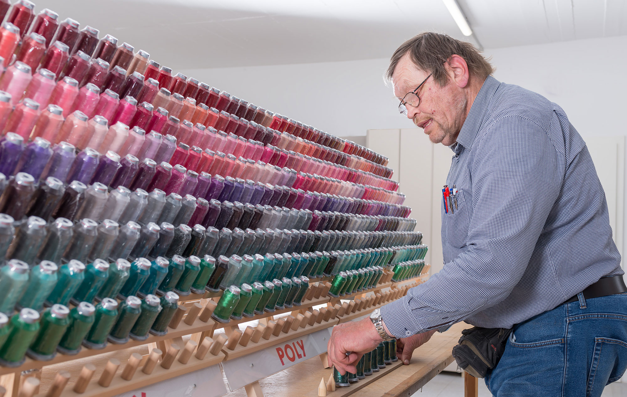 Das Bild zeigt einen Mann in einem hellblauen karierten Hemd, der sich über eine beeindruckende Sammlung von Garnspulen beugt. Die Garnspulen werden von ihm auf einem großen Holzständer systematisch nach Farben sortiert angeordnet. Die Farbpalette verläuft von oben nach unten - beginnend mit verschiedenen Rottönen über Violett und Grau bis hin zu Türkis- und Grüntönen im unteren Bereich. Die Spulen werden von ihm ordentlich in Reihen in speziellen Halterungen eingesetzt. Der Mann trägt eine Brille und setzt gerade in der vorletzten Reihe eine der Garnspulen ein. Die Szene spielt sich in einem hell ausgeleuchteten Raum mit weißer Decke ab. Das Bild zeigt einen Mann in einem hellblauen karierten Hemd, der sich über eine beeindruckende Sammlung von Garnspulen beugt. Die Garnspulen werden von ihm auf einem großen Holzständer systematisch nach Farben sortiert angeordnet. Die Farbpalette verläuft von oben nach unten - beginnend mit verschiedenen Rottönen über Violett und Grau bis hin zu Türkis- und Grüntönen im unteren Bereich. Die Spulen werden von ihm ordentlich in Reihen in speziellen Halterungen eingesetzt. Der Mann trägt eine Brille und setzt gerade in der vorletzten Reihe eine der Garnspulen ein. Die Szene spielt sich in einem hell ausgeleuchteten Raum mit weißer Decke ab.