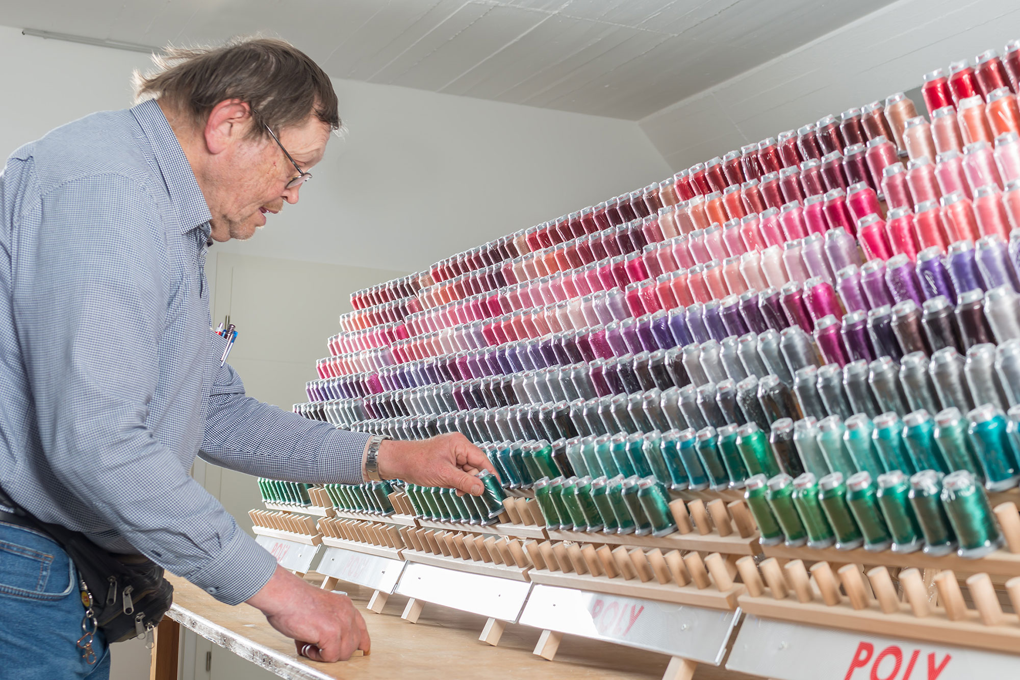 Das Bild zeigt einen Mann in einem hellblauen karierten Hemd, der sich über eine beeindruckende Sammlung von Garnspulen beugt. Die Garnspulen werden von ihm auf einem großen Holzständer systematisch nach Farben sortiert angeordnet. Die Farbpalette verläuft von oben nach unten - beginnend mit verschiedenen Rottönen über Violett und Grau bis hin zu Türkis- und Grüntönen im unteren Bereich. Die Spulen sind ordentlich in Reihen in speziellen Halterungen eingesetzt. Am unteren Rand des Regals ist teilweise die Aufschrift "POLY" zu erkennen, was auf Polyestergarn hinweist. Der Mann trägt eine Brille und setzt gerade in der vorletzten reihe eine der Garnspulen ein. Die Szene spielt sich in einem hell ausgeleuchteten Raum mit weißer Decke ab. Das Bild zeigt einen Mann in einem hellblauen karierten Hemd, der sich über eine beeindruckende Sammlung von Garnspulen beugt. Die Garnspulen werden von ihm auf einem großen Holzständer systematisch nach Farben sortiert angeordnet. Die Farbpalette verläuft von oben nach unten - beginnend mit verschiedenen Rottönen über Violett und Grau bis hin zu Türkis- und Grüntönen im unteren Bereich. Die Spulen sind ordentlich in Reihen in speziellen Halterungen eingesetzt. Am unteren Rand des Regals ist teilweise die Aufschrift "POLY" zu erkennen, was auf Polyestergarn hinweist. Der Mann trägt eine Brille und setzt gerade in der vorletzten reihe eine der Garnspulen ein. Die Szene spielt sich in einem hell ausgeleuchteten Raum mit weißer Decke ab.