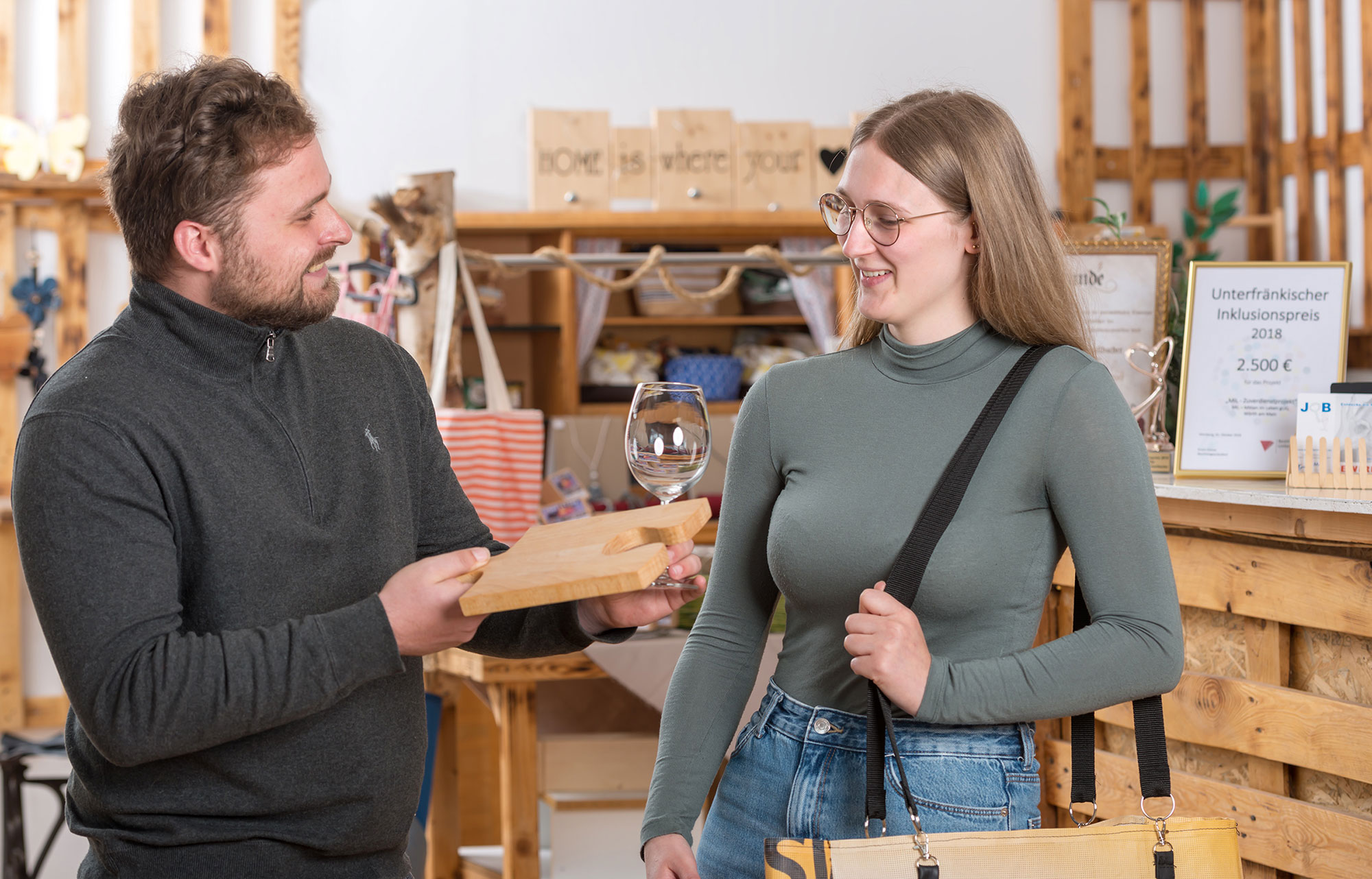 facettenReich Kundengespräch Das Bild zeigt den Verkaufsbereich des Geschäfts facettenReich. Zu sehen ist eine Theke aus Holzpaletten. Auf der Theke stehen kleinere Dekorationsartikel. Im Hintergrund sind an der Wand Holzregale mit bunten Blumen- und Herz-Dekorationen angebracht. Rechts hängt ein rotes Schild mit dem Spruch: „Wer den Tag mit einem Lachen beginnt, hat ihn bereits gewonnen.“ Im Vordergrund ist ein weißer Birkenstamm zu sehen, an dem mehrere Taschen aus Fahrradschlauch aufgehängt sind.