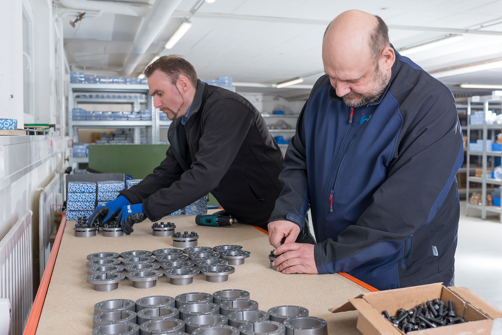Außeneinsatz - Metallbearbeitung Das Bild zeigt eine Werkstattumgebung mit zwei Personen, die gemeinsam an einem Arbeitstisch stehen. Auf dem Tisch befinden sich zahlreiche metallische Bauteile, die kreisförmig angeordnet sind, sowie mehrere blaue Verpackungen mit einem Muster aus weißen Kreisen. Eine der Personen bearbeitet die Bauteile mit einem Akku-Schrauber. Die andere Person steht daneben und bearbeitet fügt zwei Bauteile mit Schrauben zusammen. Im Hintergrund sind Regale mit weiteren Bauteilen und Verpackungen zu sehen, die ordentlich gestapelt sind. Die Szene vermittelt den Eindruck einer präzisen und strukturierten Arbeitsweise in einer industriellen oder handwerklichen Umgebung.