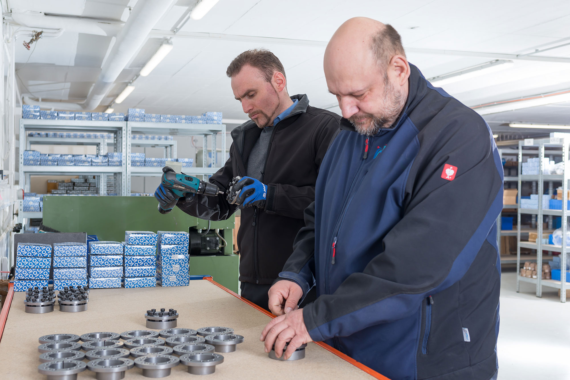 Das Bild zeigt eine Werkstattumgebung mit zwei Personen, die gemeinsam an einem Arbeitstisch stehen. Auf dem Tisch befinden sich zahlreiche metallische Bauteile, die kreisförmig angeordnet sind, sowie mehrere blaue Verpackungen mit einem Muster aus weißen Kreisen. Eine der Personen hält ein elektrisches Werkzeug, vermutlich einen Akkuschrauber, und scheint an einem der Bauteile zu arbeiten. Die andere Person steht daneben und bearbeitet ebenfalls ein Bauteil. Im Hintergrund sind Regale mit weiteren Bauteilen und Verpackungen zu sehen, die ordentlich gestapelt sind. Die Szene vermittelt den Eindruck einer präzisen und strukturierten Arbeitsweise in einer industriellen oder handwerklichen Umgebung. Das Bild zeigt eine Werkstattumgebung mit zwei Personen, die gemeinsam an einem Arbeitstisch stehen. Auf dem Tisch befinden sich zahlreiche metallische Bauteile, die kreisförmig angeordnet sind, sowie mehrere blaue Verpackungen mit einem Muster aus weißen Kreisen. Eine der Personen hält ein elektrisches Werkzeug, vermutlich einen Akkuschrauber, und scheint an einem der Bauteile zu arbeiten. Die andere Person steht daneben und bearbeitet ebenfalls ein Bauteil. Im Hintergrund sind Regale mit weiteren Bauteilen und Verpackungen zu sehen, die ordentlich gestapelt sind. Die Szene vermittelt den Eindruck einer präzisen und strukturierten Arbeitsweise in einer industriellen oder handwerklichen Umgebung.