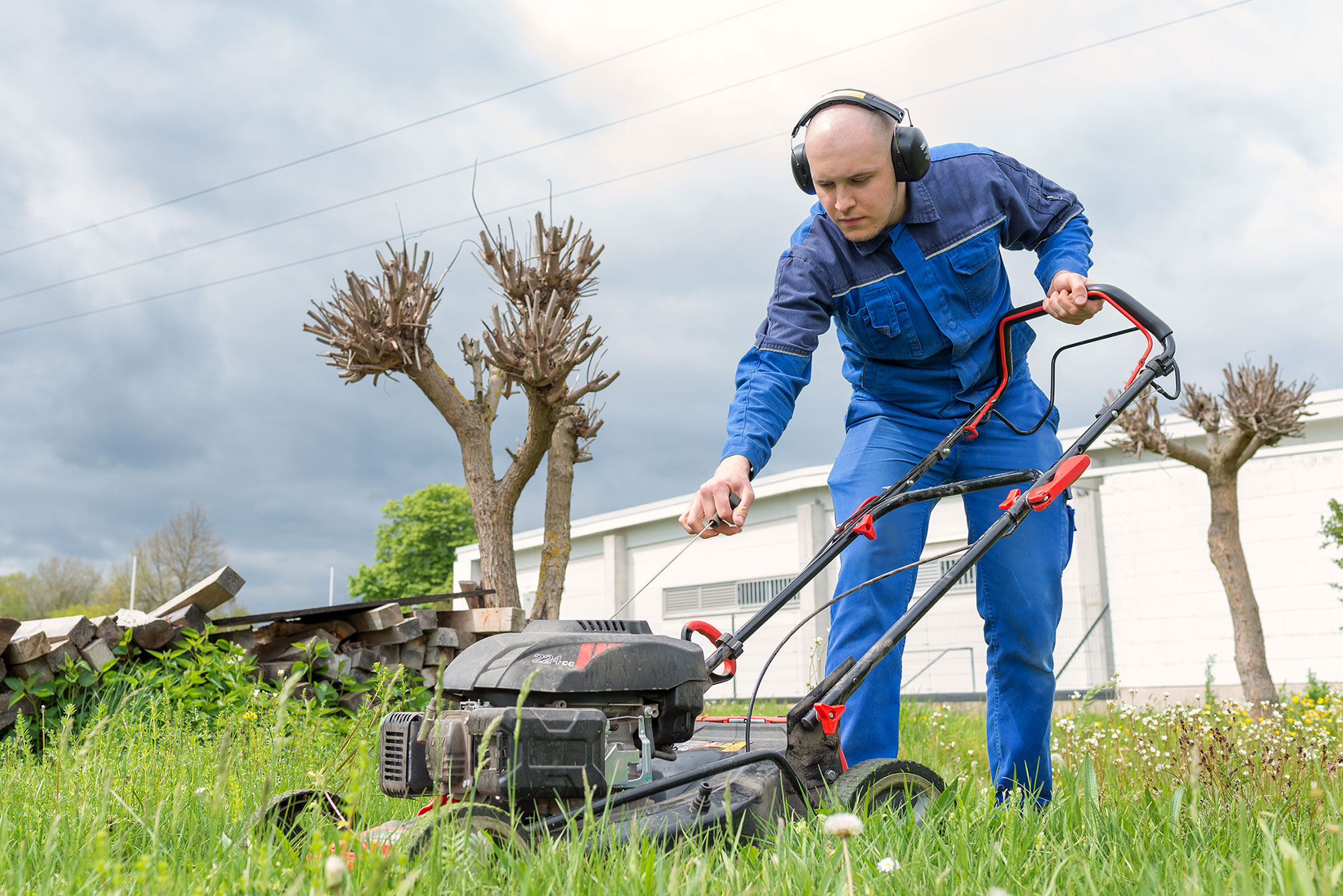Gartenpflege Das Bild zeigt einen Mann bei der Gartenarbeit. Er trägt einen blauen Arbeitsanzug und Gehörschutz während er einen Rasenmäher bedient. Der Mann arbeitet konzentriert auf einer Wiese mit hohem Gras und Wildblumen. Im Hintergrund sind beschnittene Bäume zu sehen, die typisch für einen gepflegten Außenbereich sind. Der Himmel ist leicht bewölkt und im Hintergrund ist ein weißes Gebäude zu erkennen. Der verwendete Rasenmäher scheint ein modernes Modell mit roten Akzenten zu sein. Die Szene vermittelt den Eindruck einer professionellen Grünflächenpflege.