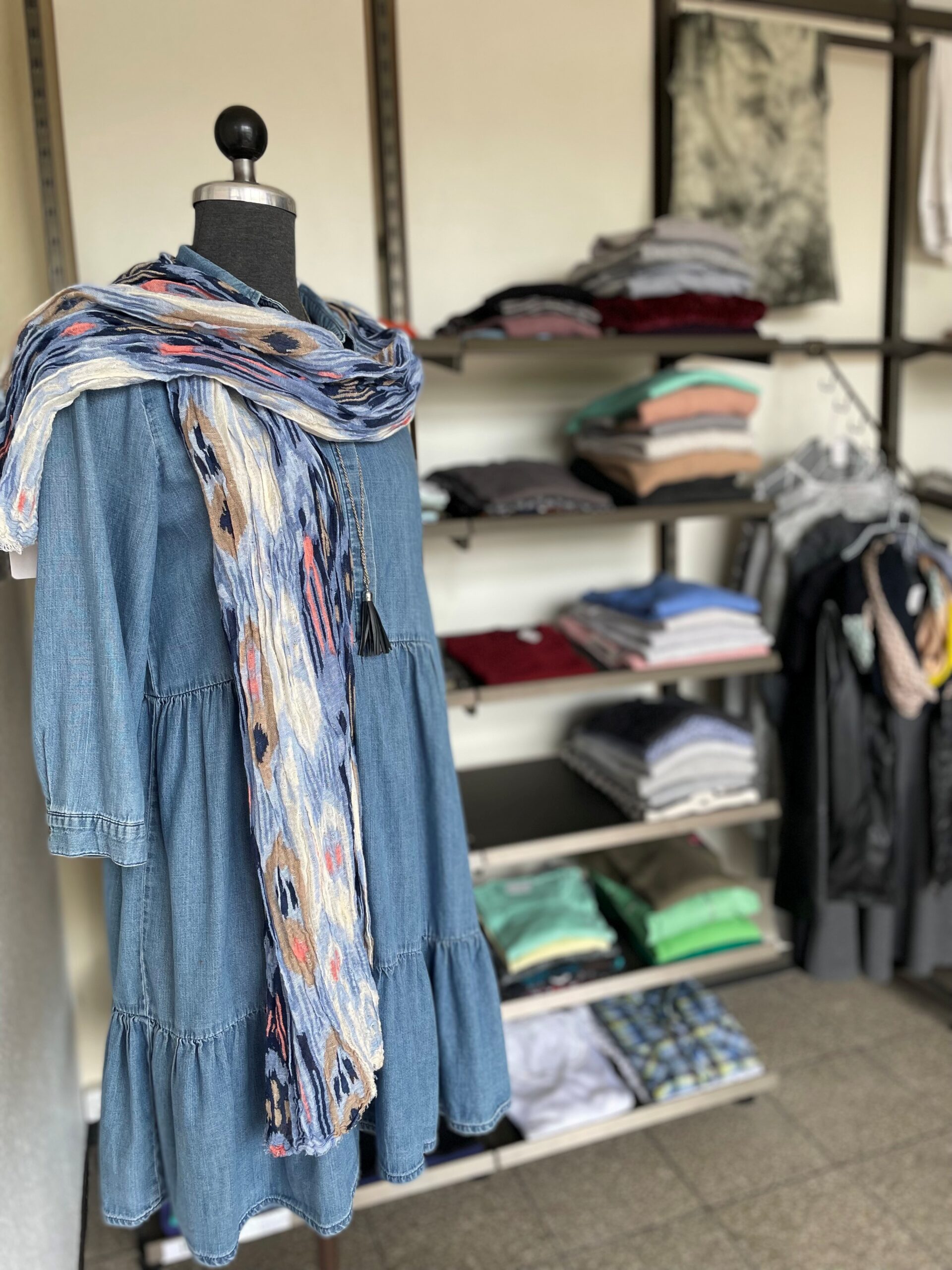 Kleiderei Das Bild zeigt eine Schaufensterpuppe, die ein blaues Jeanskleid trägt. Um den Hals der Puppe ist ein gemusterter Schal drapiert, der mit blauen, weißen und orangefarbenen Akzenten gestaltet ist. Die Puppe steht vor einem Regal, das mit gefalteter Kleidung in verschiedenen Farben und Materialien bestückt ist. Im Hintergrund sind weitere Kleidungsstücke auf Kleiderbügeln sowie dekorative Stoffe an der Wand zu sehen. Der Boden besteht aus grauen Fliesen, und die Szene vermittelt einen Eindruck vom Angebot des Second-Hand-Shops „Kleiderei“.
