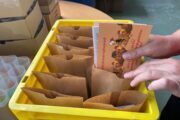 Das Bild zeigt eine gelbe Kunststoffkiste, die mit mehreren braunen Papiertüten gefüllt ist. Eine Hand steckt gerade mehrere Postkarten in eine Tüte. Links neben der Kiste stehen durchsichtige Plastikbecher mit bunten Inhalten, die anschließend ebenfalls in die Tüte konfektioniert werden müssen. Im Hintergrund sind Kartons auf einem Holztisch zu sehen.