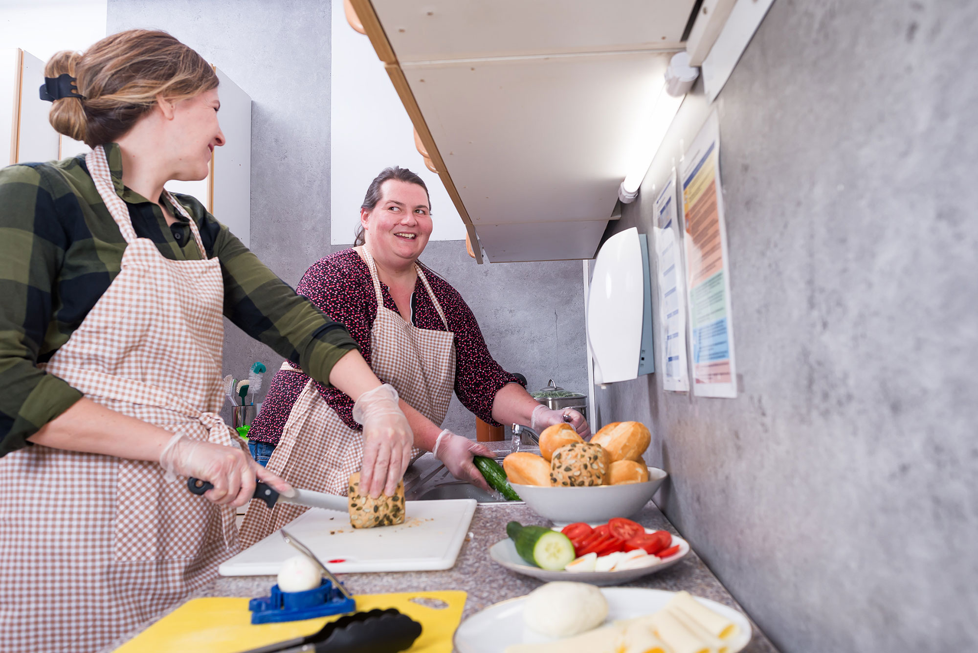 Hauswirtschaft Das Bild zeigt eine Küchenszene, in der zwei Personen gemeinsam Essen zubereiten. Sie tragen beide karierte Schürzen und Einweghandschuhe. Im Vordergrund sieht man verschiedene Lebensmittel auf der Arbeitsfläche: Geschnittenes Gemüse wie Gurken und Tomaten auf einem Teller. Mehrere Brötchen in einer Schale, darunter einige, die aussehen wie Körnerbrötchen sowie ein Schneidebrett, auf dem gerade etwas geschnitten wird. Die Küche hat eine graue Wand und ist mit Lampen beleuchtet. An der Wand hängen einige Informationsblätter oder Poster. Die Atmosphäre wirkt entspannt und freundlich, da beide Personen lächeln und in ihre Arbeit vertieft sind. Die Situation findet im Hauswirtschaftsbereich von MiL statt, worauf die professionelle Einrichtung und die Hygienemaßnahmen (Handschuhe) hindeuten.