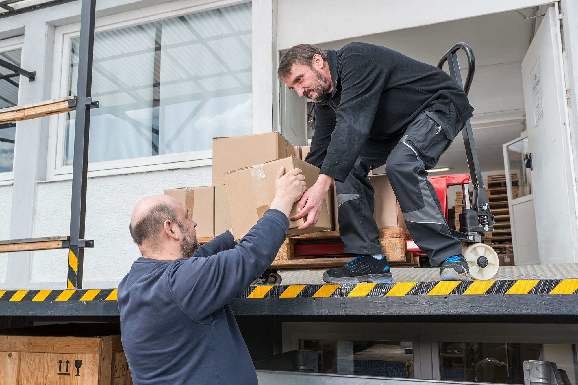 Lager und Logistik Das Bild zeigt eine Verladesituation an einer Laderampe. Zwei Männer arbeiten zusammen beim Verladen von Kartons. Ein Mann steht auf der erhöhten Laderampe, die mit einem schwarz-gelben Warnstreifen markiert ist, während der andere Mann von unten die Kartons anreicht. Der Mann auf der Rampe trägt schwarze Arbeitskleidung mit grauen Elementen und blaue Sicherheitsschuhe. Sein Kollege trägt einen dunkelblauen Pullover. Im Hintergrund sind ein weißes Gebäude mit Fenstern und einige Holzpaletten zu sehen. Die Szene vermittelt einen professionellen Eindruck von Teamarbeit bei logistischen Tätigkeiten.