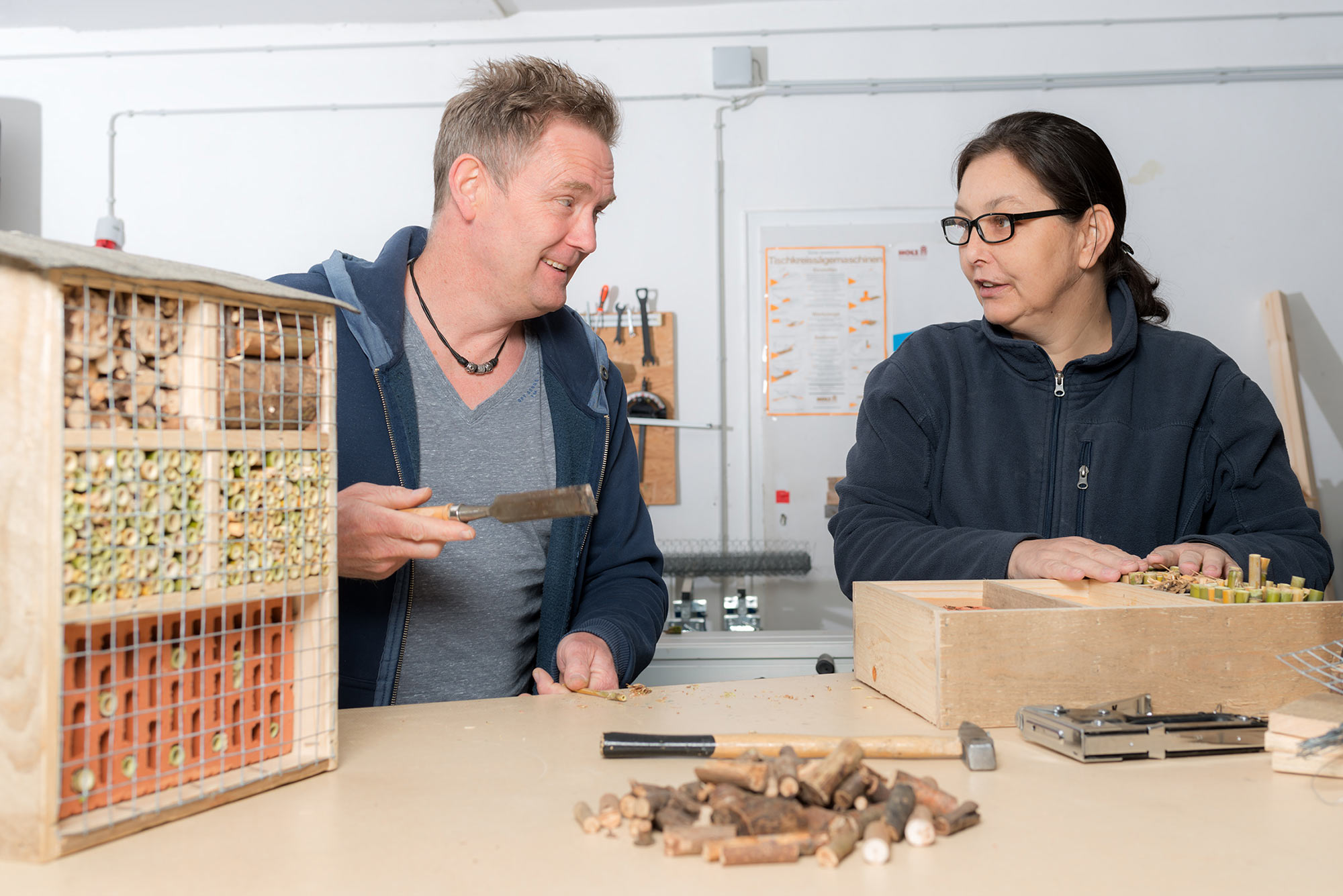 Holzwerkstatt Das Bild zeigt zwei Personen in der therapeutischen Werkstatt, die an einem Holzprojekt arbeiten. Sie stehen an einem Arbeitstisch, auf dem verschiedene Holzstücke und Werkzeuge verteilt sind. Im Vordergrund ist ein Insektenhotel mit wabenartigen Fächern zu sehen. Die Person links trägt einen grauen V-Ausschnitt-Pullover mit einer blauen Kapuzenjacke darüber und hält ein Werkzeug in der Hand. Die andere Person trägt eine schwarze Fleecejacke und eine Brille. Beide sind in ein Gespräch vertieft und scheinen konzentriert an ihrem Projekt zu arbeiten.