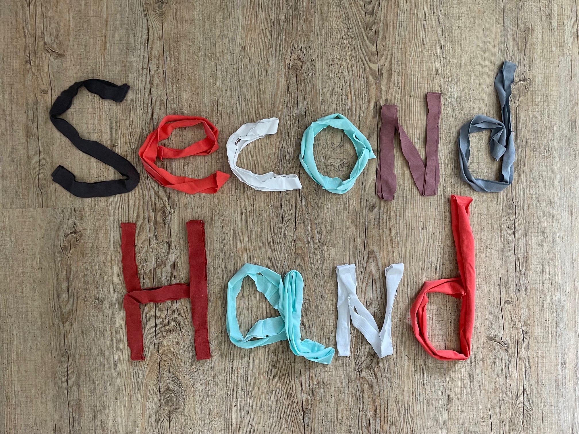 Second Hand Das Bild zeigt den Schriftzug „Second Hand“, der kreativ aus Stoffstreifen gelegt wurde. Die Buchstaben sind aus verschiedenfarbigen Stoffstreifen geformt, die auf einem Holzboden arrangiert sind. Die Farben der Stoffe variieren und umfassen Schwarz, Rot, Weiß, Hellblau, Grau und Braun. Jeder Buchstabe ist individuell gestaltet, wobei die Stoffstreifen entweder gerollt oder flach ausgelegt wurden. Der Schriftzug wirkt handgemacht und nachhaltig, passend zum Thema „Second Hand“. Die Verwendung von recycelten Stoffen unterstreicht die Botschaft von Wiederverwendung und Umweltbewusstsein.
