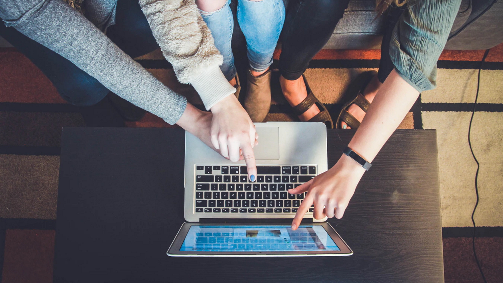 Das Bild zeigt drei Personen, die gemeinsam an einem Laptop arbeiten. Zwei von ihnen tragen Jeans, die dritte Person eine grüne Bluse. Alle drei zeigen mit ihren Fingern auf den Bildschirm des Laptops, der auf einem schwarzen Tisch steht. Der Hintergrund besteht aus einem bunten Teppich mit geometrischen Mustern. Die Szene vermittelt den Eindruck von Teamarbeit oder gemeinsamer Problemlösung. Das Bild zeigt drei Personen, die gemeinsam an einem Laptop arbeiten. Zwei von ihnen tragen Jeans, die dritte Person eine grüne Bluse. Alle drei zeigen mit ihren Fingern auf den Bildschirm des Laptops, der auf einem schwarzen Tisch steht. Der Hintergrund besteht aus einem bunten Teppich mit geometrischen Mustern. Die Szene vermittelt den Eindruck von Teamarbeit oder gemeinsamer Problemlösung.