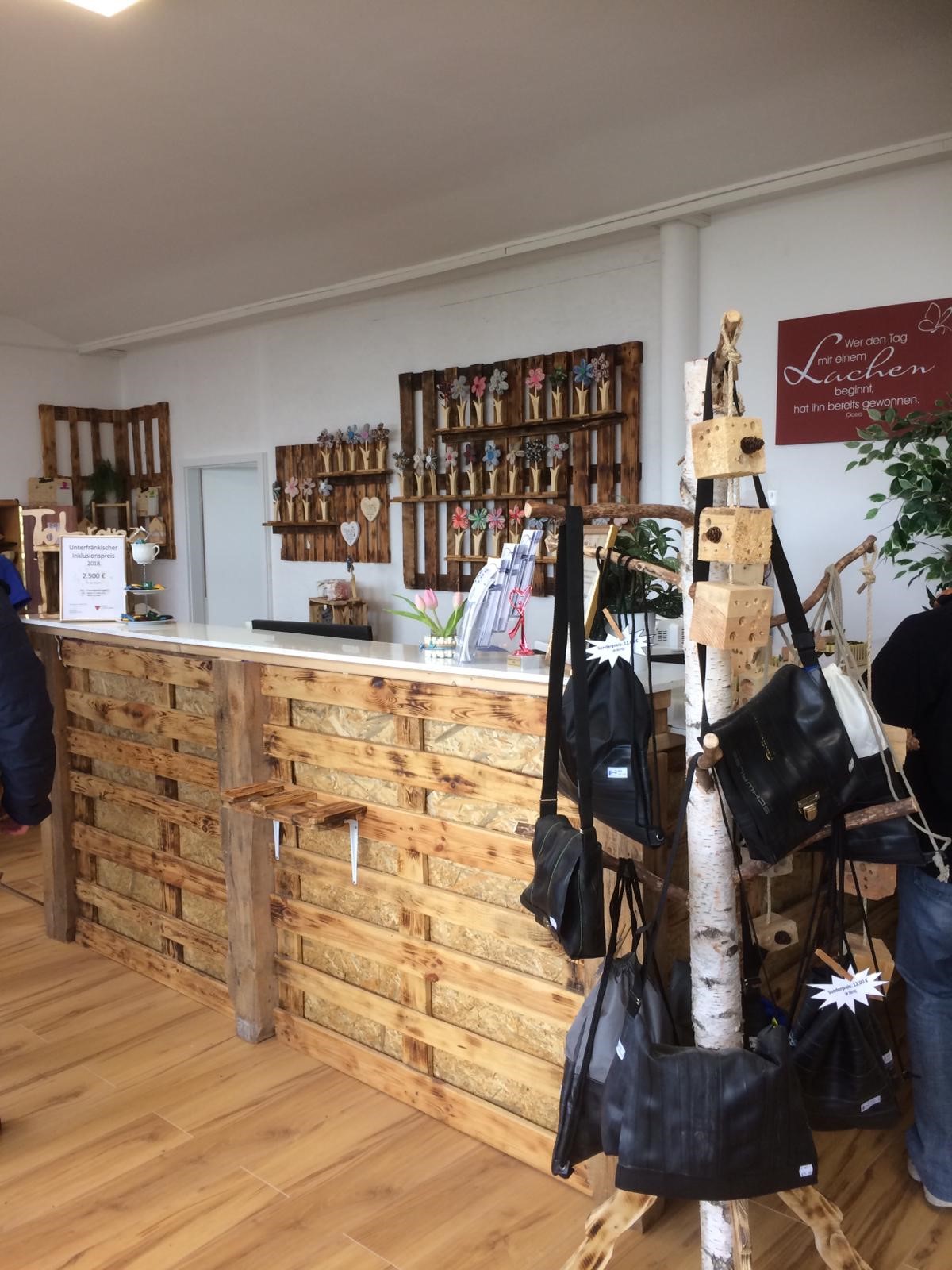 Das Bild zeigt den Verkaufsbereich des Geschäfts facettenReich. Zu sehen ist eine Theke aus Holzpaletten. Auf der Theke stehen kleinere Dekorationsartikel. Im Hintergrund sind an der Wand Holzregale mit bunten Blumen- und Herz-Dekorationen angebracht. Rechts hängt ein rotes Schild mit dem Spruch: „Wer den Tag mit einem Lachen beginnt, hat ihn bereits gewonnen.“ Im Vordergrund ist ein weißer Birkenstamm zu sehen, an dem mehrere Taschen aus Fahrradschlauch aufgehängt sind.