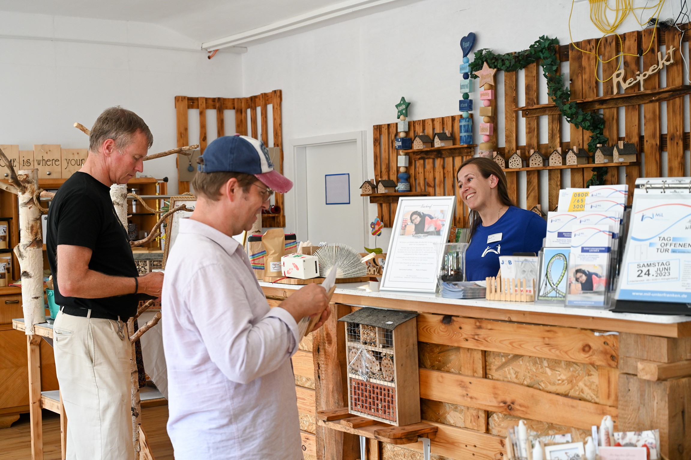 facettenReich Das Bild zeigt eine Verkaufsszene im Werkstatt-Laden facettenReich. Der Raum ist mit Holzpaletten an den Wänden gestaltet und dekorativ eingerichtet. Zwei Kunden - einer in schwarzem T-Shirt und einer in weißem Hemd mit Baseballkappe - betrachten Waren, während eine Mitarbeiterin in blauem Shirt freundlich hinter der Verkaufstheke steht. Auf der Theke sind verschiedene Broschüren und Werbematerialien ausgelegt. An den Wänden befinden sich kleine Holzhäuschen, Sterne und grüne Ranken als Dekoration.
