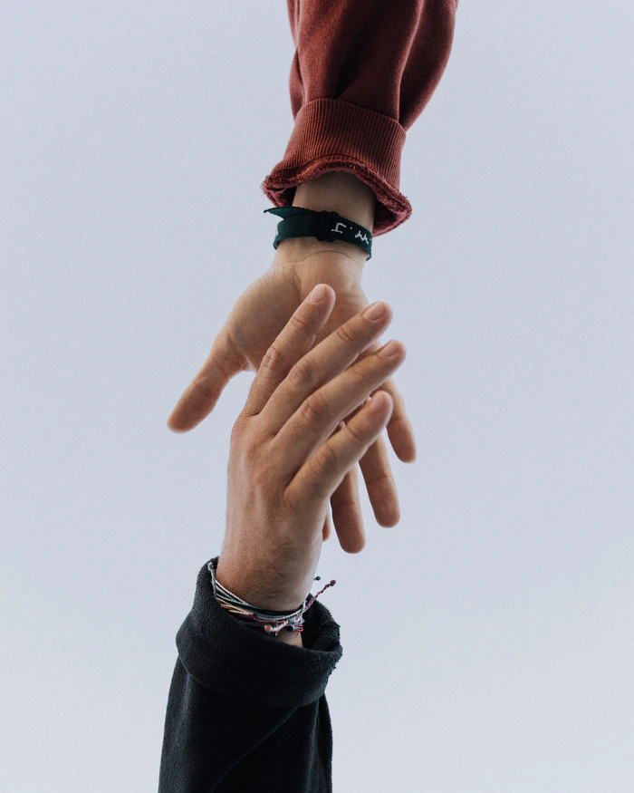 Das Bild zeigt zwei Hände, die sich berühren. Sie sollen helfende Hände symbolisieren. Die obere Hand gehört einer Person mit einem roten Pullover und trägt ein schwarzes Armband mit einem weißen Muster. Die untere Hand gehört einer Person mit einem schwarzen Kleidungsstück und mehreren bunten Armbändern aus dünnen Schnüren. Der Hintergrund ist schlicht und hellgrau, wodurch die Hände im Fokus stehen. Das Bild zeigt zwei Hände, die sich berühren. Sie sollen helfende Hände symbolisieren. Die obere Hand gehört einer Person mit einem roten Pullover und trägt ein schwarzes Armband mit einem weißen Muster. Die untere Hand gehört einer Person mit einem schwarzen Kleidungsstück und mehreren bunten Armbändern aus dünnen Schnüren. Der Hintergrund ist schlicht und hellgrau, wodurch die Hände im Fokus stehen.