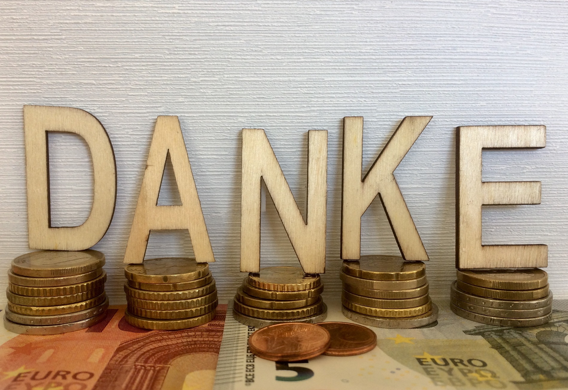 Das Bild zeigt das Wort „DANKE“, das aus einzelnen Holzbuchstaben besteht. Jeder Buchstabe steht auf einem Stapel aus Euro-Münzen, die in verschiedenen Größen und Werten angeordnet sind. Im Vordergrund liegen zwei Kupfermünzen, während im Hintergrund ein 50-Euro-Schein sichtbar ist. Der Hintergrund besteht aus einer weißen Wand mit einer feinen, strukturierten Oberfläche. Das Bild zeigt das Wort „DANKE“, das aus einzelnen Holzbuchstaben besteht. Jeder Buchstabe steht auf einem Stapel aus Euro-Münzen, die in verschiedenen Größen und Werten angeordnet sind. Im Vordergrund liegen zwei Kupfermünzen, während im Hintergrund ein 50-Euro-Schein sichtbar ist. Der Hintergrund besteht aus einer weißen Wand mit einer feinen, strukturierten Oberfläche.