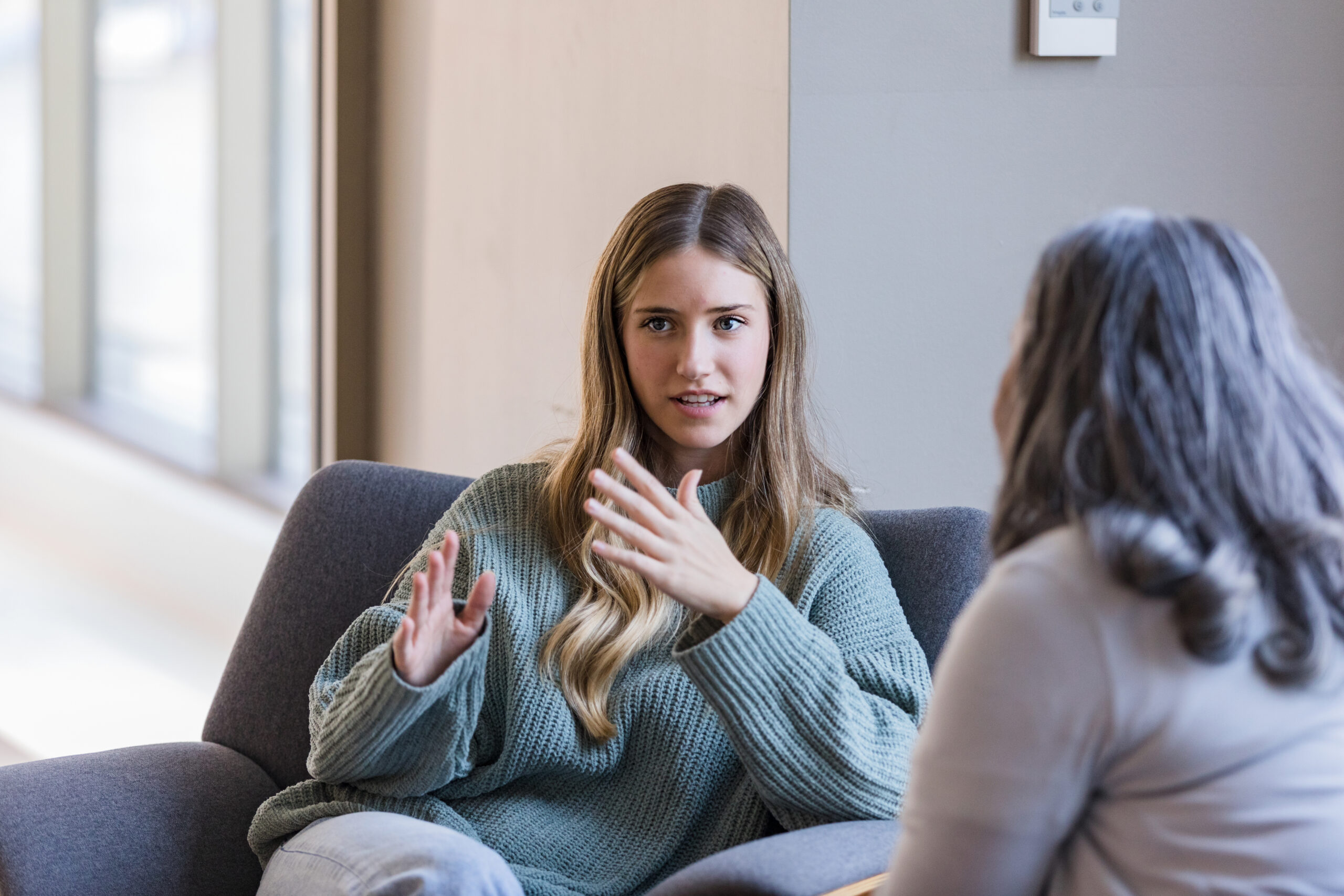 Im Gespräch Zwei Frauen sitzen sich in einem hellen Raum auf bequemen Sesseln gegenüber. Die junge Frau im Vordergrund trägt einen grob gestrickten, hellgrünen Pullover. Sie spricht und macht mit den Händen Gesten. Sie schaut die andere Frau nicht an. Die andere Frau ist von hinten zu sehen und hat schulterlanges, graues Haar. Im Hintergrund sind große Fenster und eine helle Wand. Die Szene wirkt ruhig und aufmerksam. Das Bild zeigt, wie wichtig es ist, zuzuhören, wenn Menschen mit psychischen Erkrankungen schwierige Zeiten durchmachen.