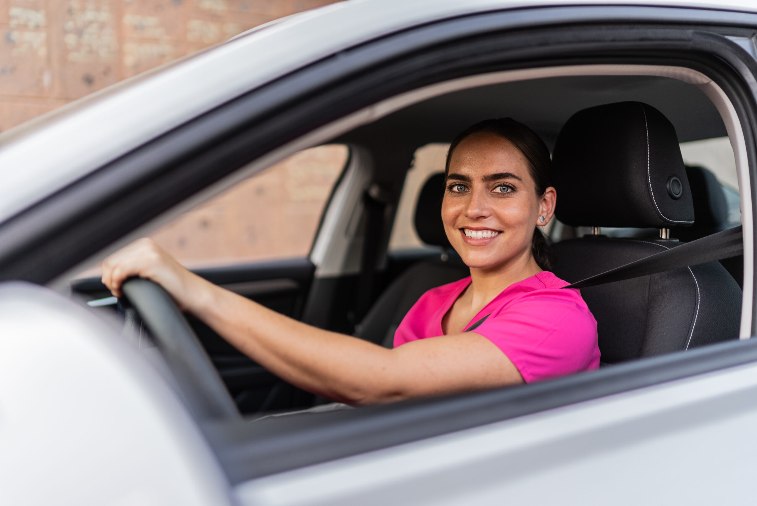 Unterwegs zum Hausbesuch im ABW Eine junge Frau sitzt auf dem Fahrersitz eines Autos. Sie trägt ein pinkfarbenes Oberteil. Sie hat den Sicherheitsgurt angelegt. Mit einer Hand hält sie das Lenkrad. Das Autofenster ist offen. Im Hintergrund ist eine beige Wand zu sehen, die unscharf ist. Das Bild wurde von außen durch das offene Fenster aufgenommen. Es zeigt eine Fahrt zu einem Hausbesuch im Rahmen des Ambulant Betreuten Wohnens.