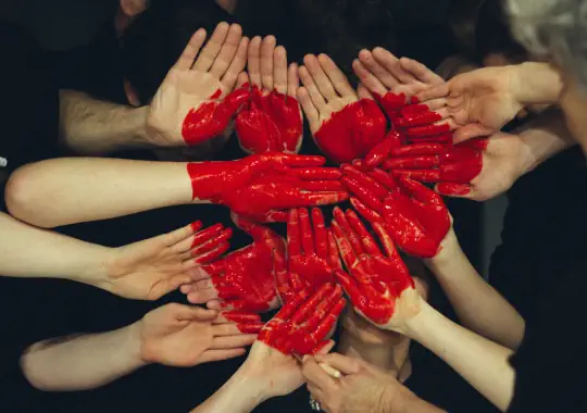 Viele Hände - rotes Herz Das Bild zeigt eine künstlerische Aufnahme von mehreren Händen, die in einem Kreis angeordnet sind. Die Handflächen sind mit roter Farbe bemalt und bilden zusammen die Form eines Herzens. Die intensive rote Farbe steht in starkem Kontrast zum dunklen Hintergrund, was dem Bild eine besonders ausdrucksstarke und symbolische Wirkung verleiht. Diese Komposition vermittelt ein Gefühl von Gemeinschaft, Zusammenhalt und geteilter Emotionen.