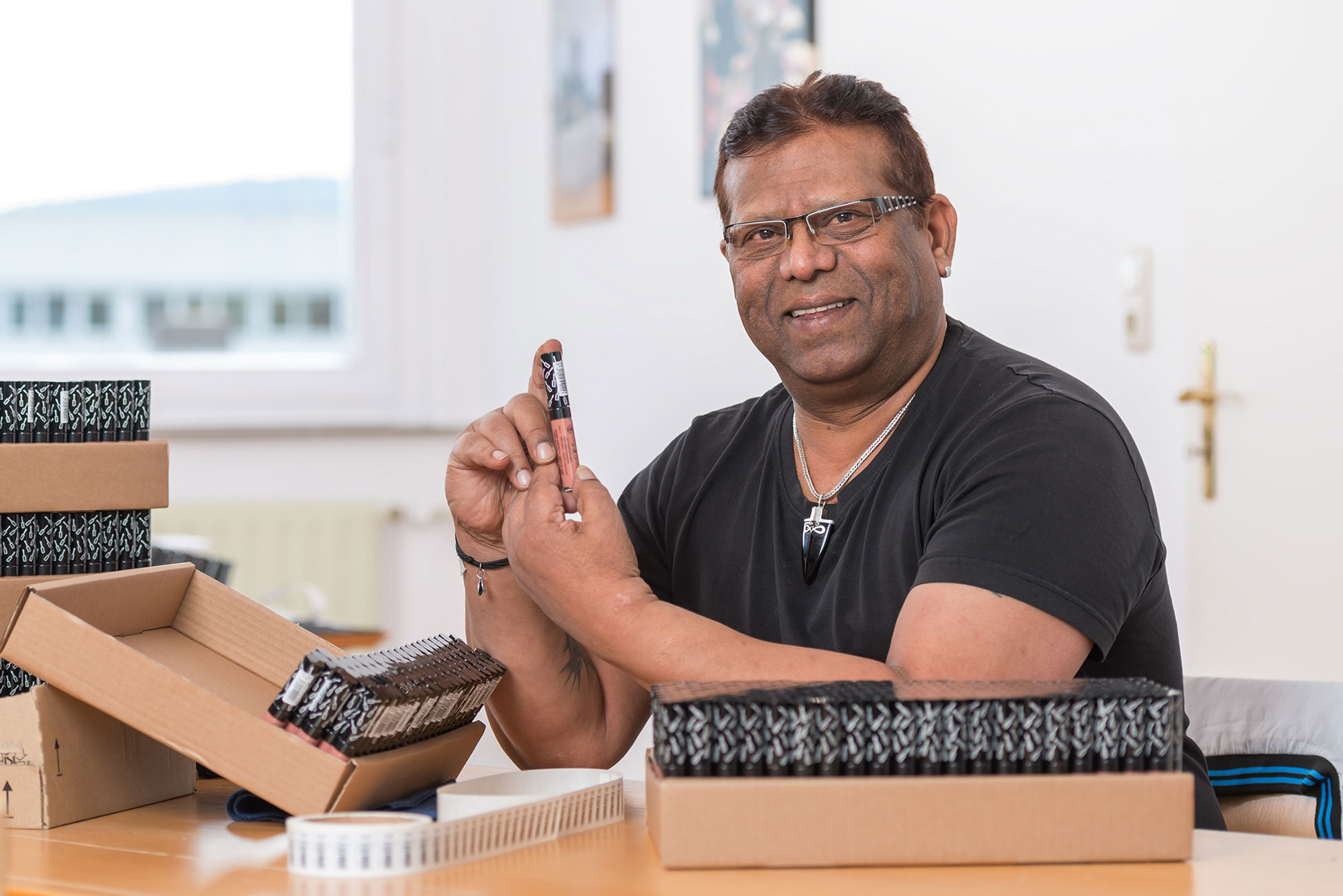 230426_MiL_Etikettierung_022_web Das Bild zeigt einen Mann mittleren Alters in einem schwarzen T-Shirt, der konzentriert ein kleines Objekt in seinen Händen etikettiert. Er trägt eine Brille und eine silberne Halskette mit einem Anhänger. Vor ihm auf dem Tisch befinden sich mehrere braune Kartons und Boxen mit weiteren Artikeln. Der Raum, in dem er sich befindet, ist hell und minimalistisch eingerichtet, mit weißen Wänden und einem Fenster im Hintergrund. Es handelt sich um einen Arbeitsplatz im Zuverdienstprojekt, an dem sorgfältig Firmenaufträge bearbeitet werden.